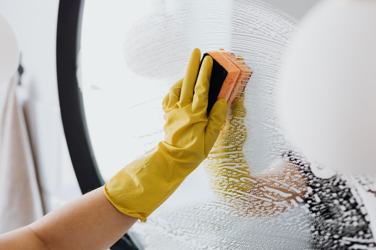 Yellow-gloved hand scrubbing a mirror with a sponge